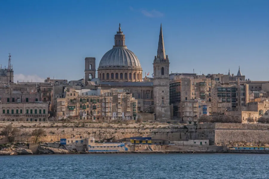 Valletta cityscape from the water, featuring a large dome, a tall spire, and historic buildings along a rocky shore.