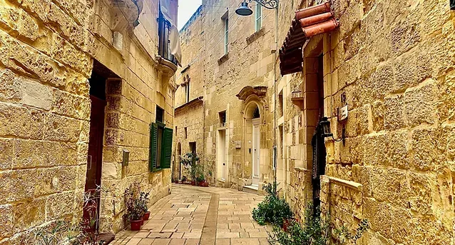 A narrow stone street in an old town, flanked by tall buildings with balconies, green shutters, and ornate street lamps.
