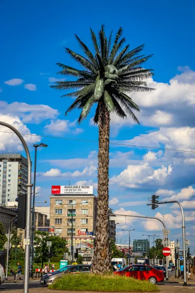 Tall artificial palm tree on a grassy island in a city intersection with buildings, traffic, and pedestrians under a blue ...