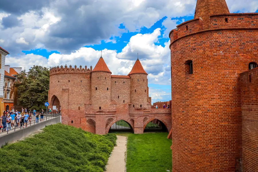 Red brick barbican with two conical towers and arched passages. Green grass and a path lead up to it, with people walking ...
