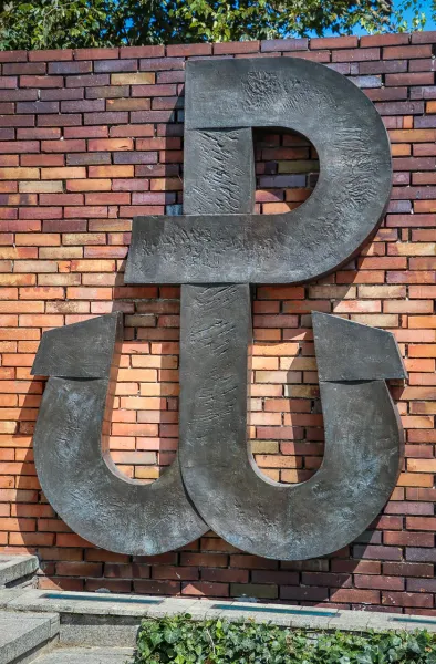 Large, dark metal sculpture of the Fighting Poland (Kotwica) symbol on a multi-colored brick wall with green leaves above ...