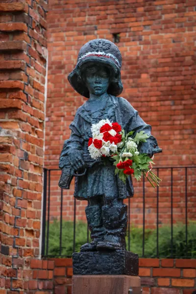 Bronze statue of a child soldier in a helmet holding red and white flowers, against a brick wall.
