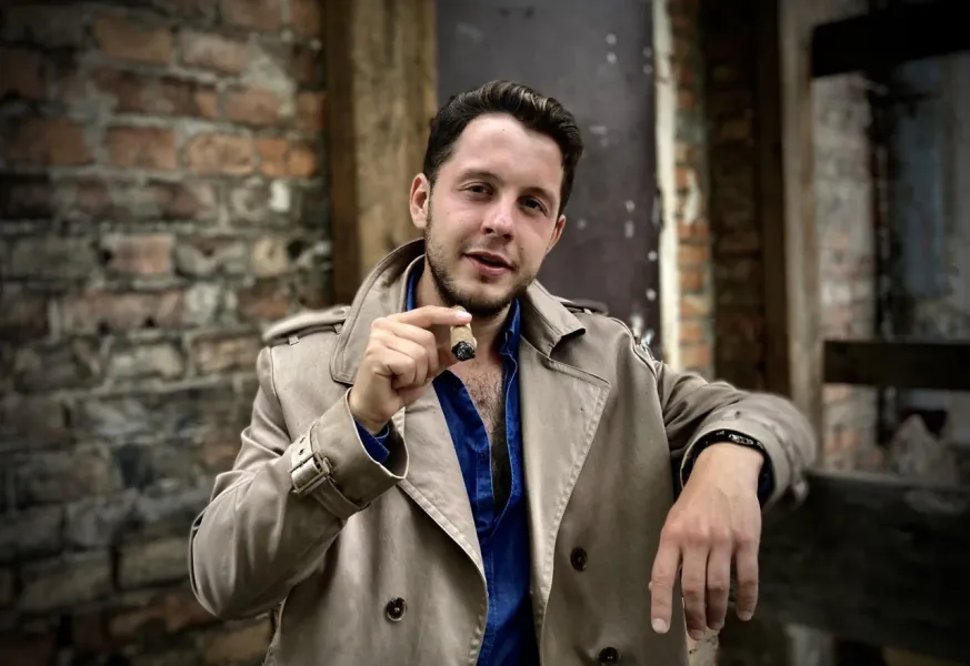 Young man in a beige trench coat and blue shirt holding a lit cigar, standing in front of an old brick wall.