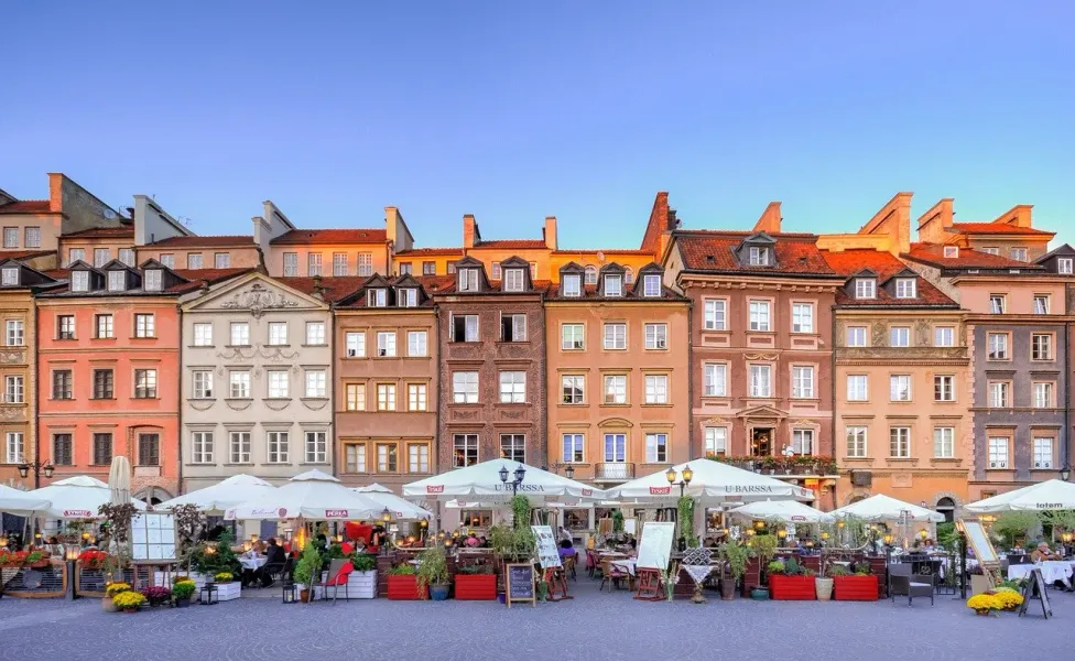 Colorful historic buildings with red roofs overlook an outdoor cafe area with white umbrellas and people dining under a bl...
