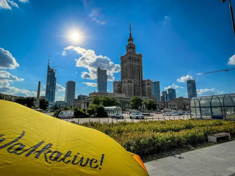 Palace of Culture and Science and modern skyscrapers under a sunny blue sky, with a yellow Walkative umbrella in the foreg...