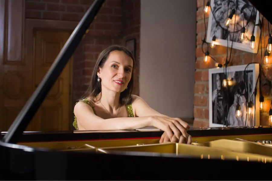 Woman in green top smiling, resting hands on a black grand piano. Brick wall and string lights in the background.