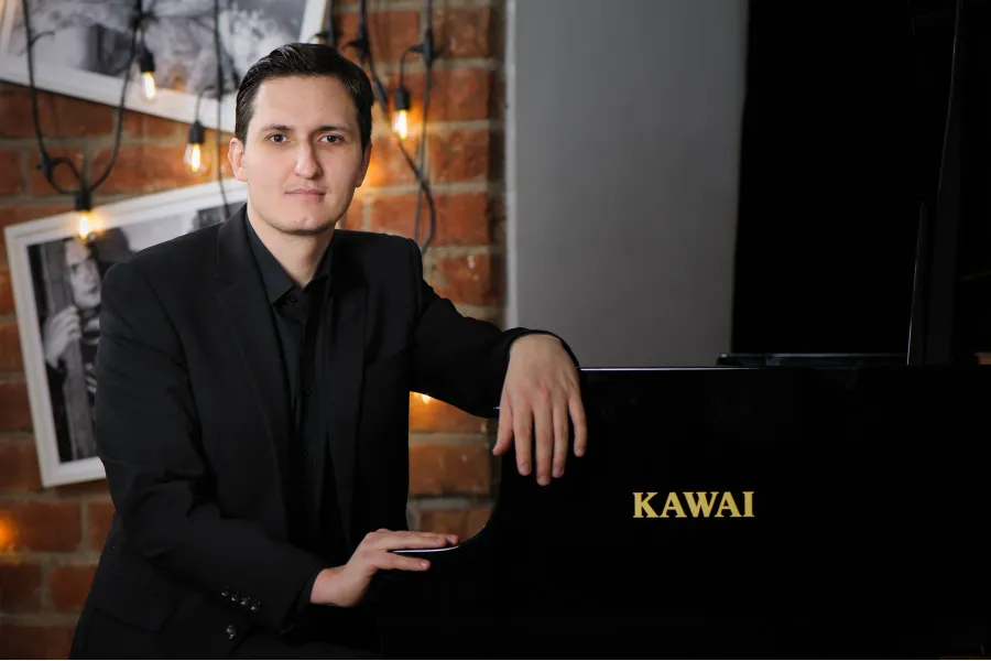 Man in a black suit leaning on a black Kawai piano in a room with a brick wall and string lights in Warsaw.
