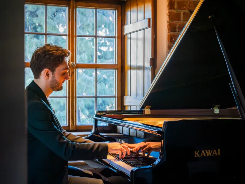 Man in a suit playing a black Kawai grand piano next to a window with wooden shutters.