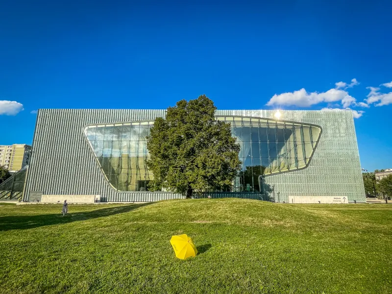 Modern museum building with a wavy glass facade and metal panels, a large green tree, and a yellow umbrella on a grassy mo...