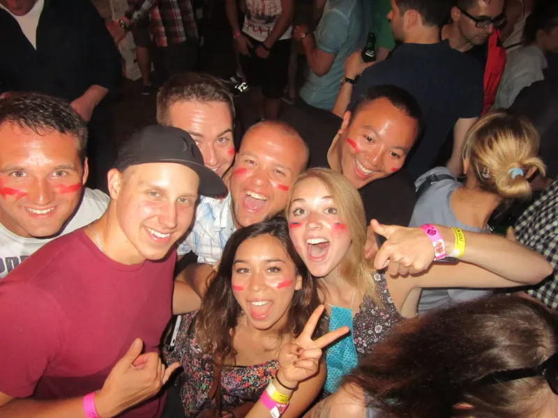 A diverse group of young people smiling, many with red face paint on their cheeks, posing closely together at a lively ind...