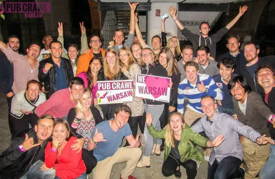 A large group of diverse young adults smiling, posing, and holding "Pub Crawl Warsaw" and "We love Warsaw" signs in an urb...