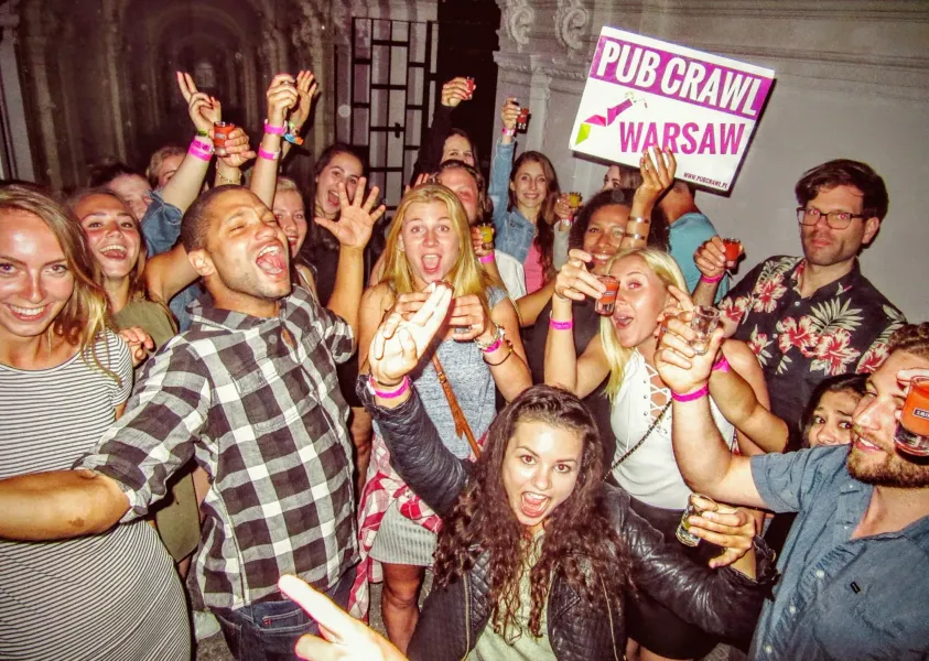 Group of young adults smiling, laughing, and holding shot glasses, one holding a 'PUB CRAWL WARSAW' sign.