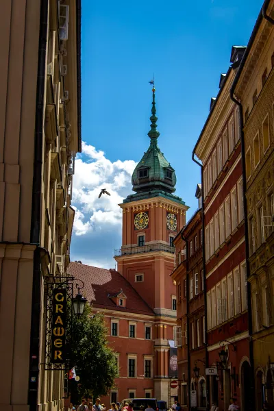 View up a narrow street towards the Royal Castle clock tower in Warsaw with a green dome and golden clock faces, a bird fl...