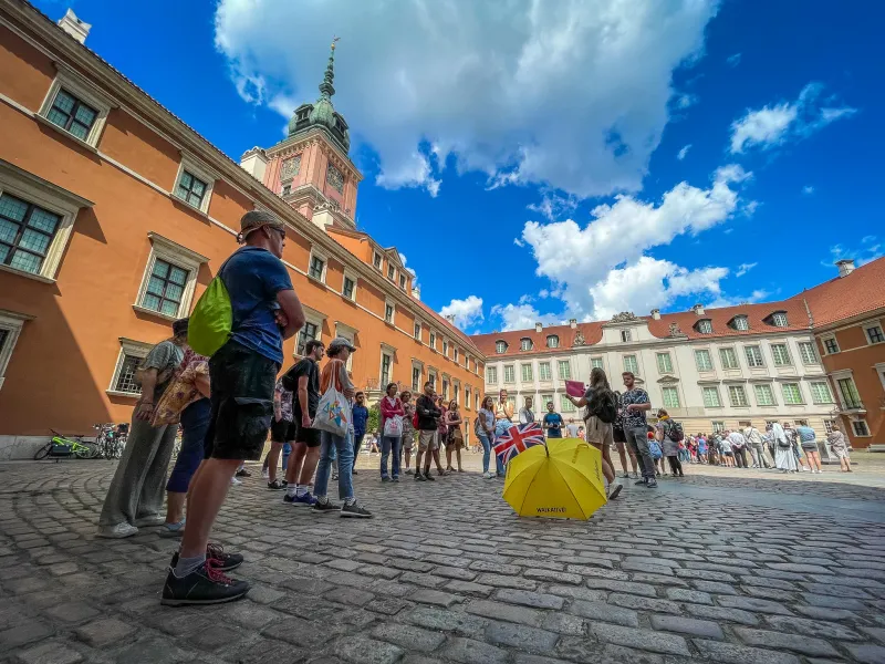 Tour group with guide and yellow umbrella on a cobblestone courtyard in front of Warsaw Royal Castle.