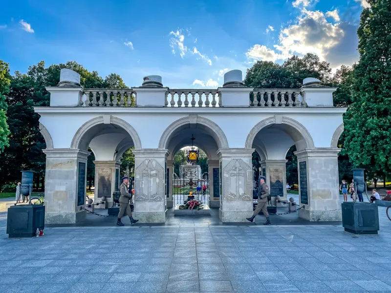 Two uniformed guards march in front of the white stone arcades of the Tomb of the Unknown Soldier in Warsaw under a blue sky.