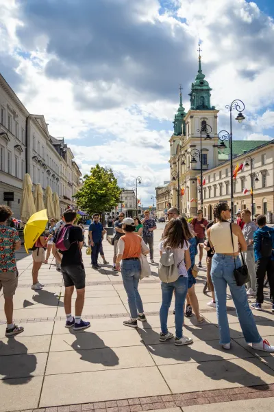 A group of people gathered on a paved street in Warsaw with a historic church and other buildings in the background under ...