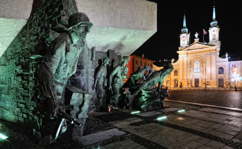 Bronze monument depicting figures of Warsaw Uprising soldiers under green light at night, with a brightly lit church behind.