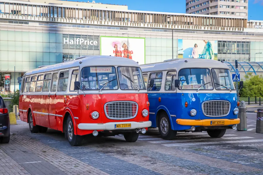 Two vintage Polish tourist buses, one red and one blue, parked on a city street in front of a modern building.