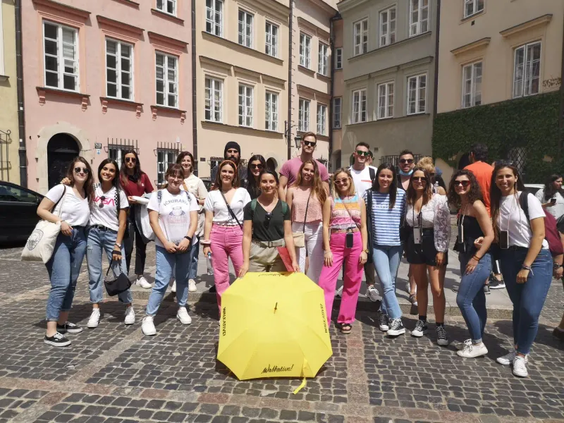 A group of young adults poses on a cobbled street with a yellow Walkative umbrella. Colorful historic buildings in backgro...
