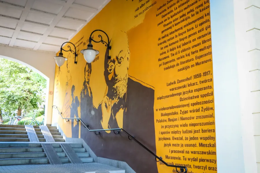 Large yellow mural depicting Ludwik Zamenhof and text, next to a concrete staircase with handrails and lamps.