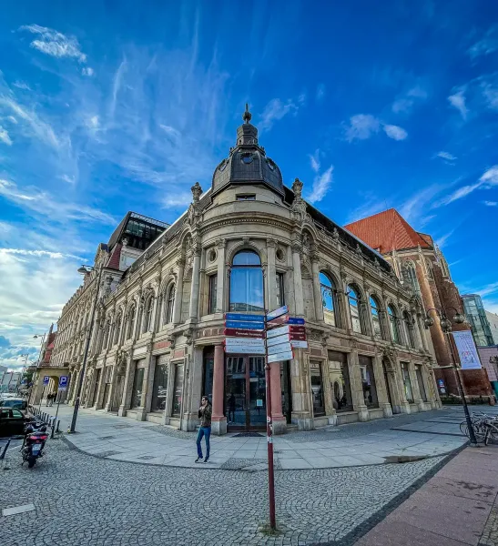 Ornate historic building with a domed corner tower and large windows on a cobblestone street in Wrocław. A street signpost...