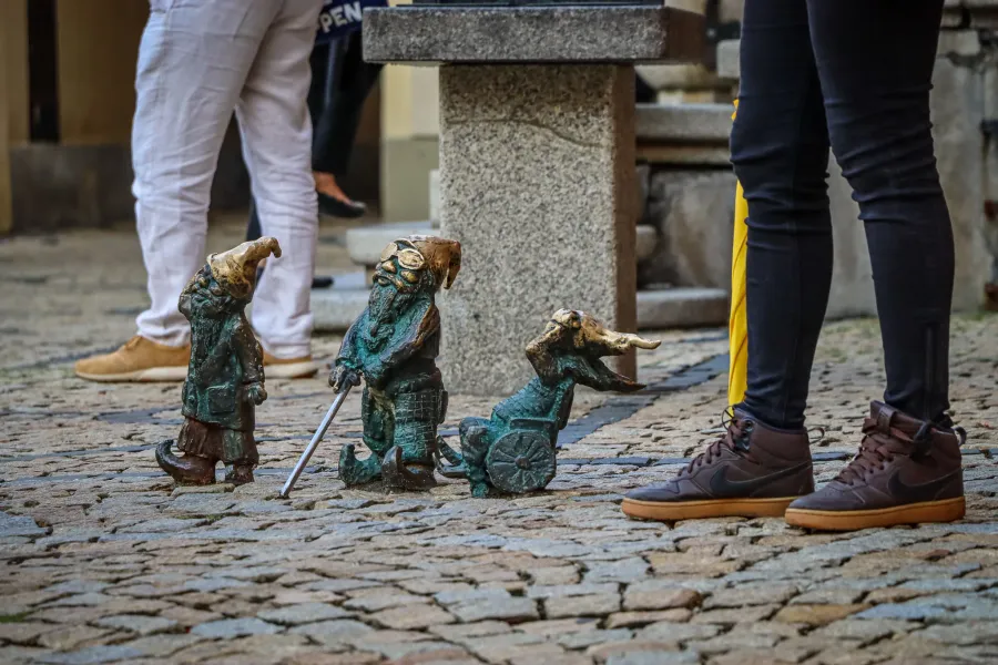 Three bronze dwarf statues on a cobblestone street, with parts of people's legs visible around them. One dwarf holds a staff.