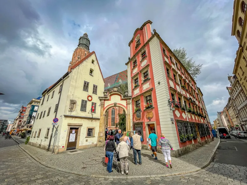 People walk on a cobblestone street in Wroclaw's Old Town, past historic yellow and red buildings, and an archway leading ...