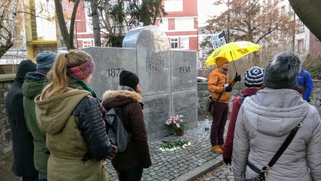 Stone monument commemorating the New Synagogue with dates 1870 and 1938, attended by a tour group with guide.