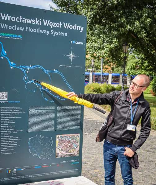 Man pointing with a yellow umbrella at an information board displaying a map of Wrocław's Floodway System on a sunny day.