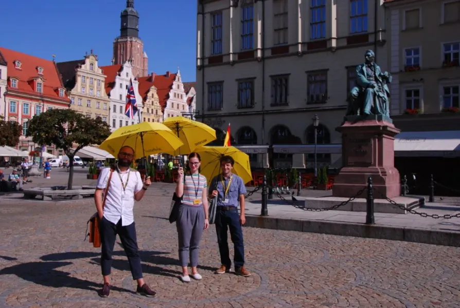 Three people with yellow umbrellas stand on a cobblestone square in front of historic buildings and a statue on a sunny day.