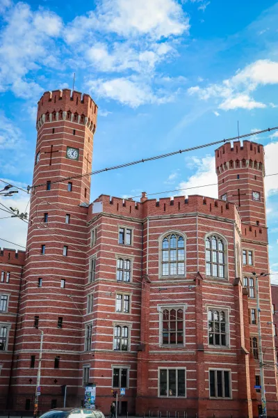 Historic red-brick building with two towers, each featuring a clock face, under a blue sky with white clouds in Wroclaw.