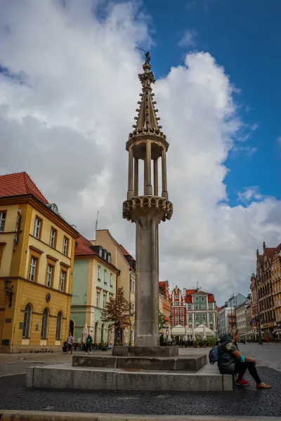 Tall stone pillory with an ornate spire and a figure on top, surrounded by colorful historic buildings in a European city ...
