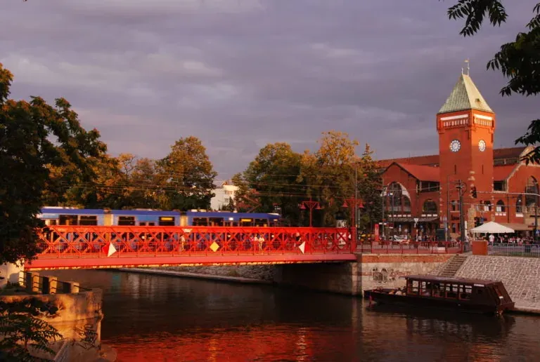 Red metal bridge over a river with a blue train crossing, next to a red brick market hall with a clock tower at sunset.