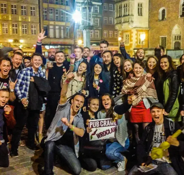 Group of cheerful people at a night pub crawl in Wrocław, with historic buildings in the background. A sign reads "Pub Cra...