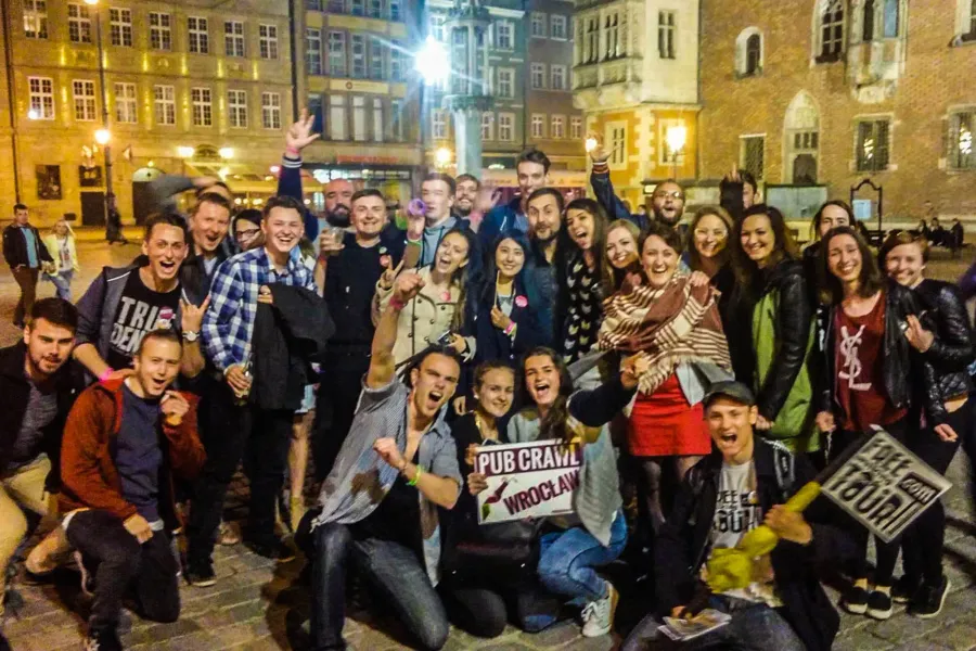 A large group of young adults smiling and posing for a photo on a street at night. One person holds a 'Pub Crawl Wrocław' ...