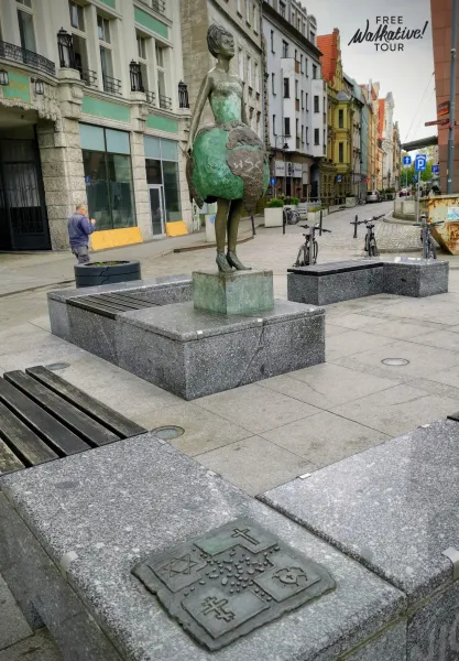 Bronze statue of a woman with a globe as a skirt, granite benches, and a metal plaque with religious symbols on a city str...