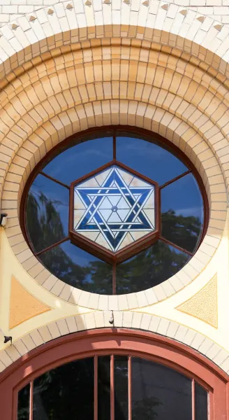 Round stained glass window with a blue and white Star of David design, set in a yellow brick arch above a brown wooden frame.