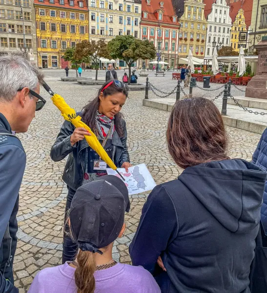Female tour guide with yellow umbrella and map explains to three tourists on a cobblestone square. Colorful historic build...