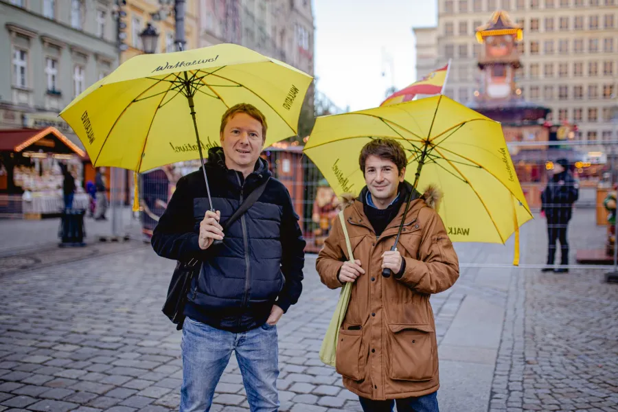 Two men holding yellow Walkative! umbrellas stand on a cobblestone square with blurred city buildings and market stalls in...