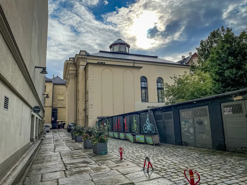 Cobblestone alleyway leading to the White Stork Synagogue in Wroclaw under a cloudy sky. Potted plants and bicycle racks a...