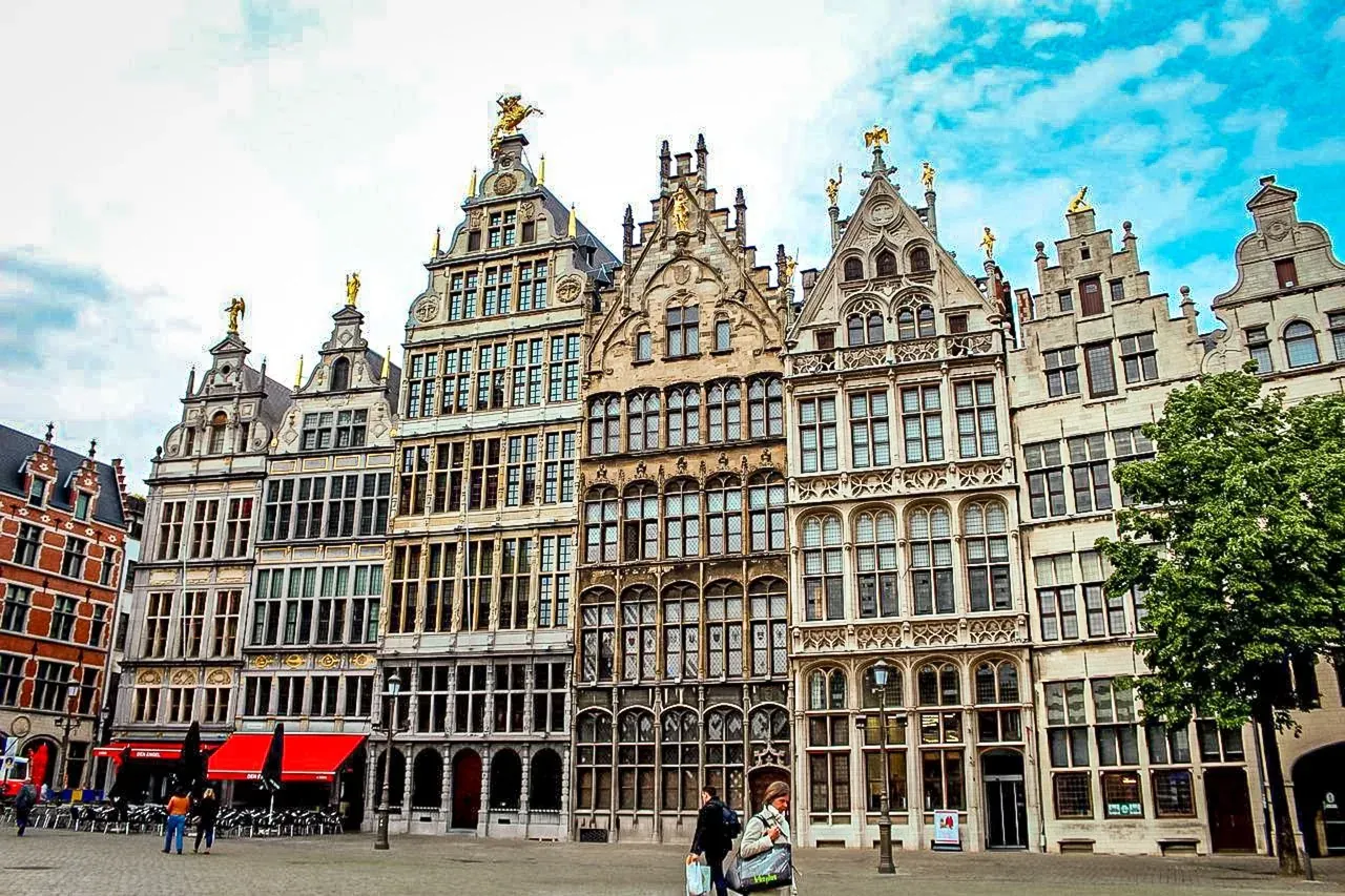 Elaborate historic guildhalls with stepped gables and golden statues line a cobblestone market square in Antwerp. People walk