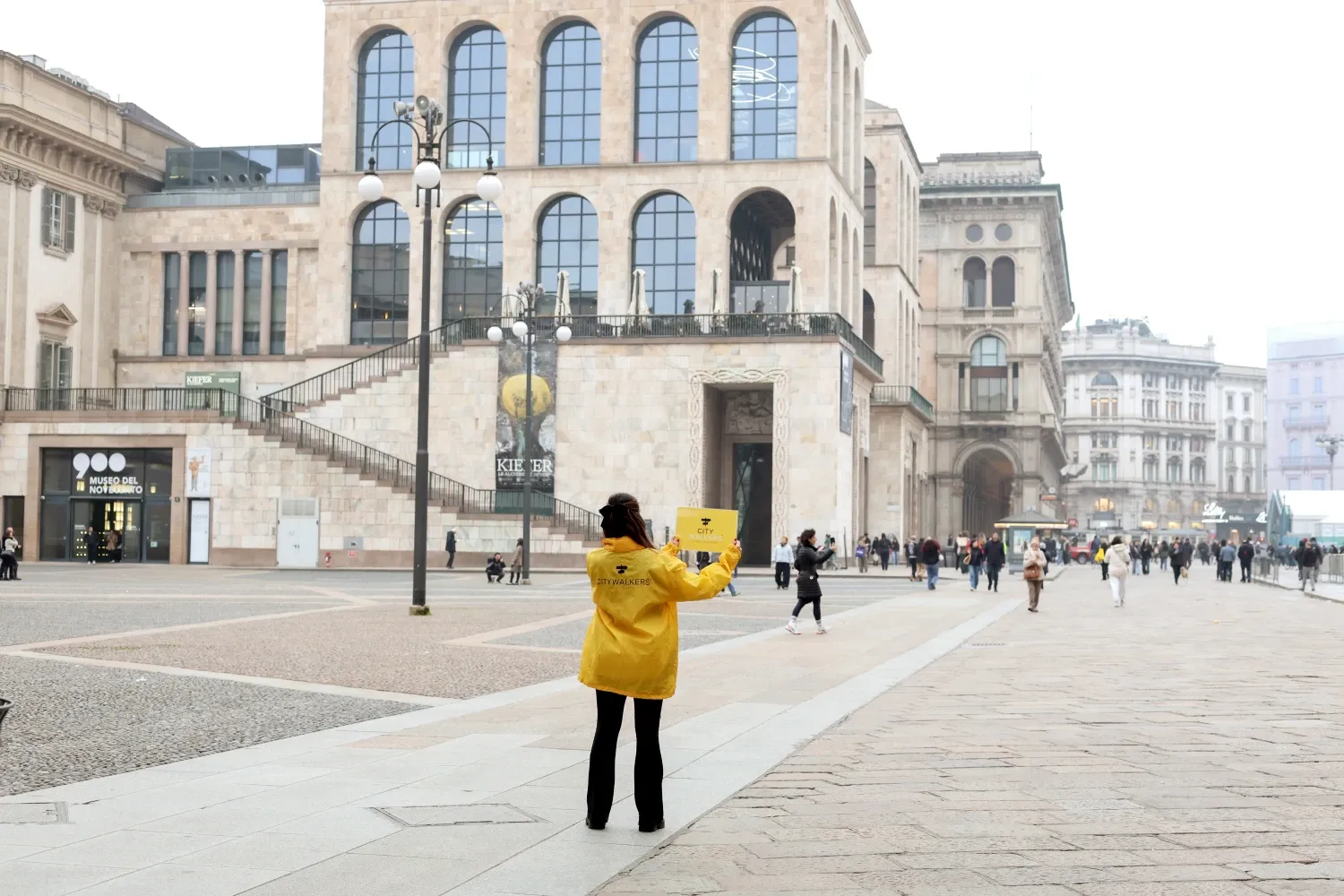 Tour guide in a yellow jacket holding a sign on Piazza del Duomo in Milan in front of the Museo del Novecento.