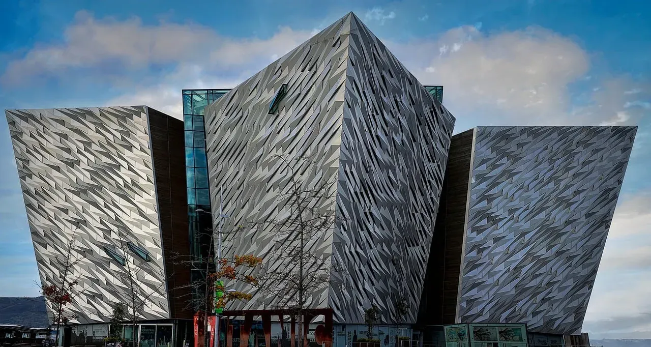 The Titanic Belfast building with its angular, faceted silver panels under a partly cloudy sky, with bare trees in the foregr