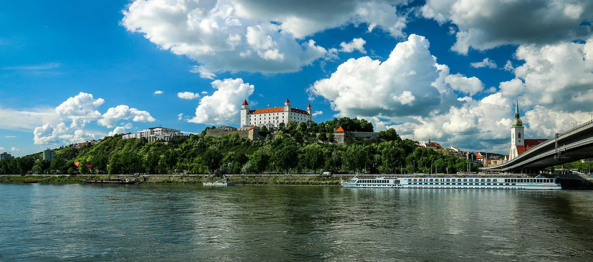 Bratislava Castle on a green hill overlooking the Danube River, with a cruise ship, bridge, and St. Martin's Cathedral spire.