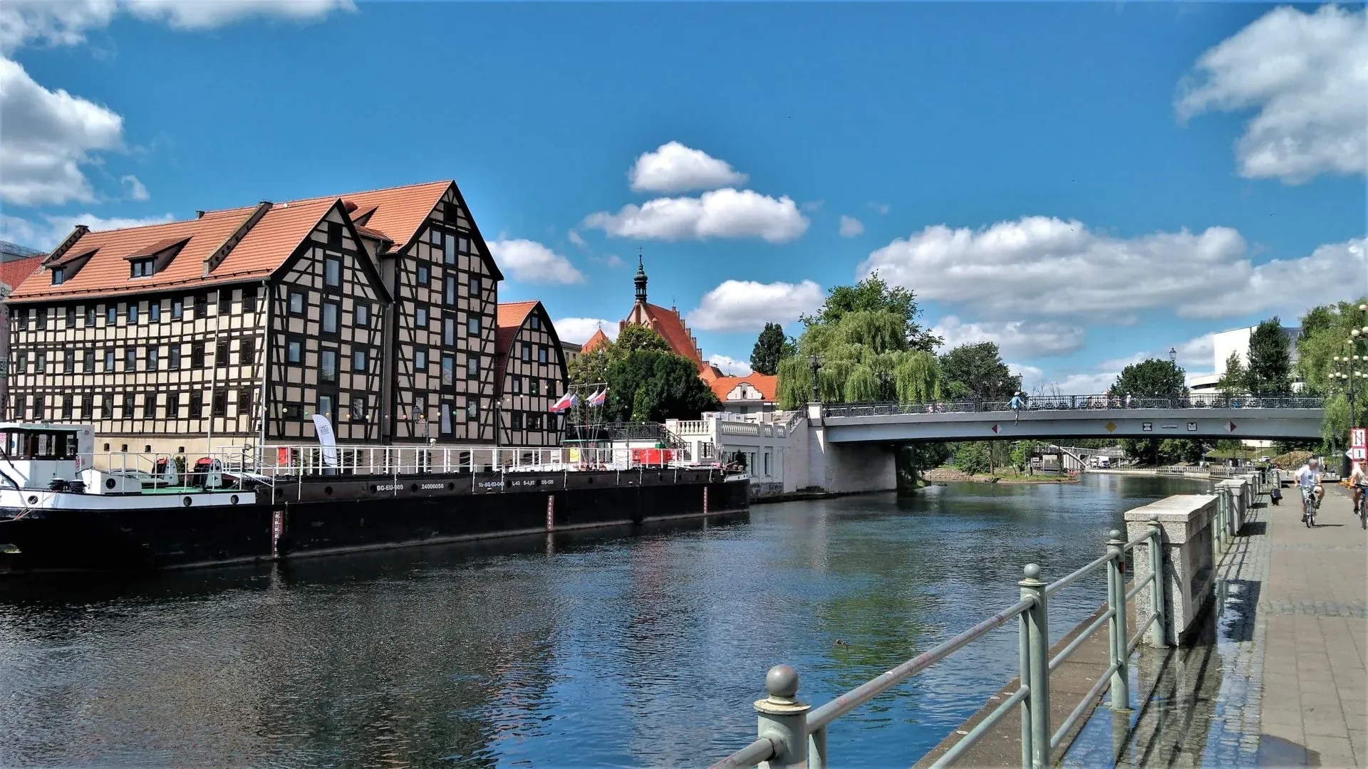 Half-timbered buildings with red roofs on the riverbank, a long barge, a concrete bridge, and a pedestrian path under a blue