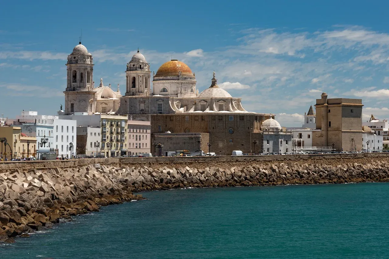View of Cadiz Cathedral and city skyline from the sea, with a rocky breakwater in the foreground under a blue sky.