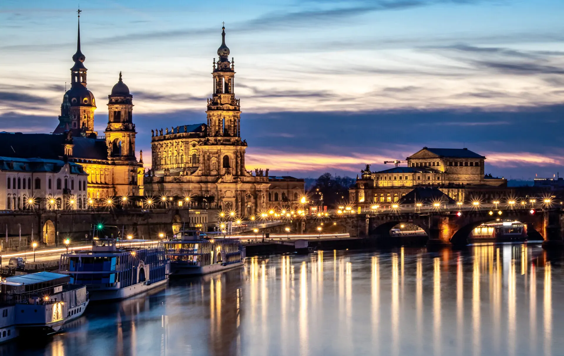 Dresden city skyline at dusk with illuminated historic buildings along the Elbe River, boats docked, and reflections on the w