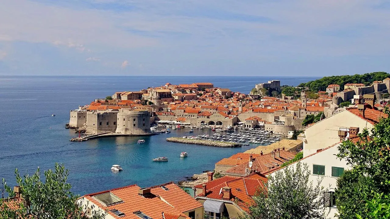 Elevated view of Dubrovnik's Old Town, featuring red-tiled roofs, medieval city walls, a harbor with boats, and the Adriatic