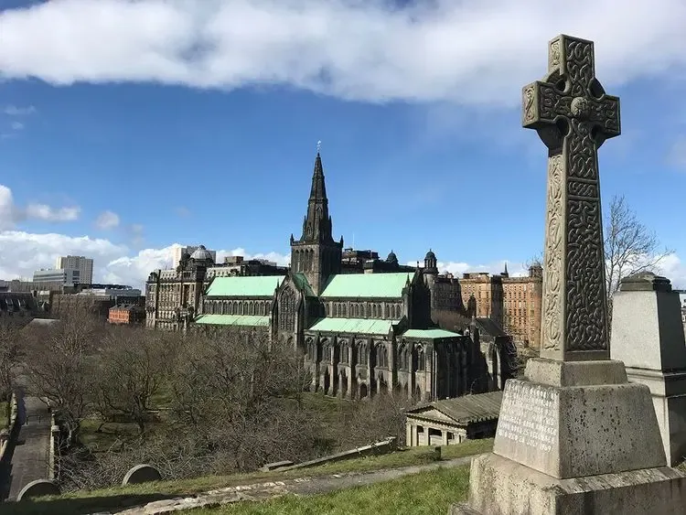 Historic Glasgow Cathedral with green roof and spire, viewed from the Necropolis, showing an ornate Celtic cross in the foreg