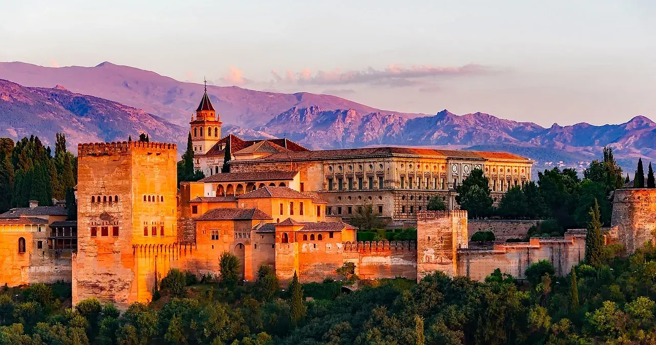Alhambra palace complex with fortified walls and towers at sunset, mountains in the background, surrounded by green trees.